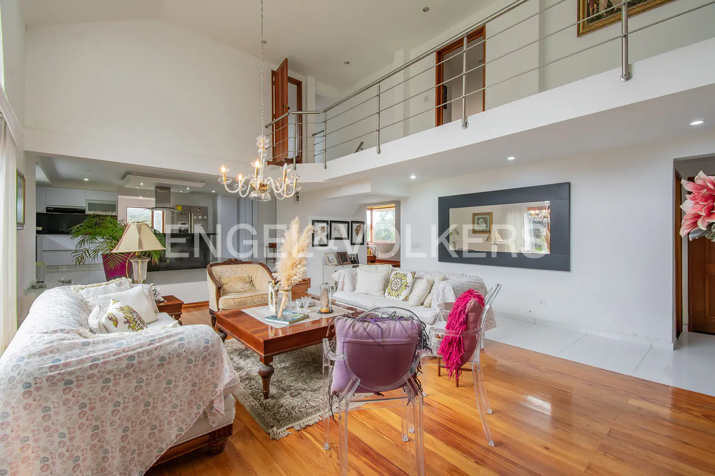 Spacious living room with high ceilings, wooden floors, a chandelier, white sofas, and a loft overlooking the area.