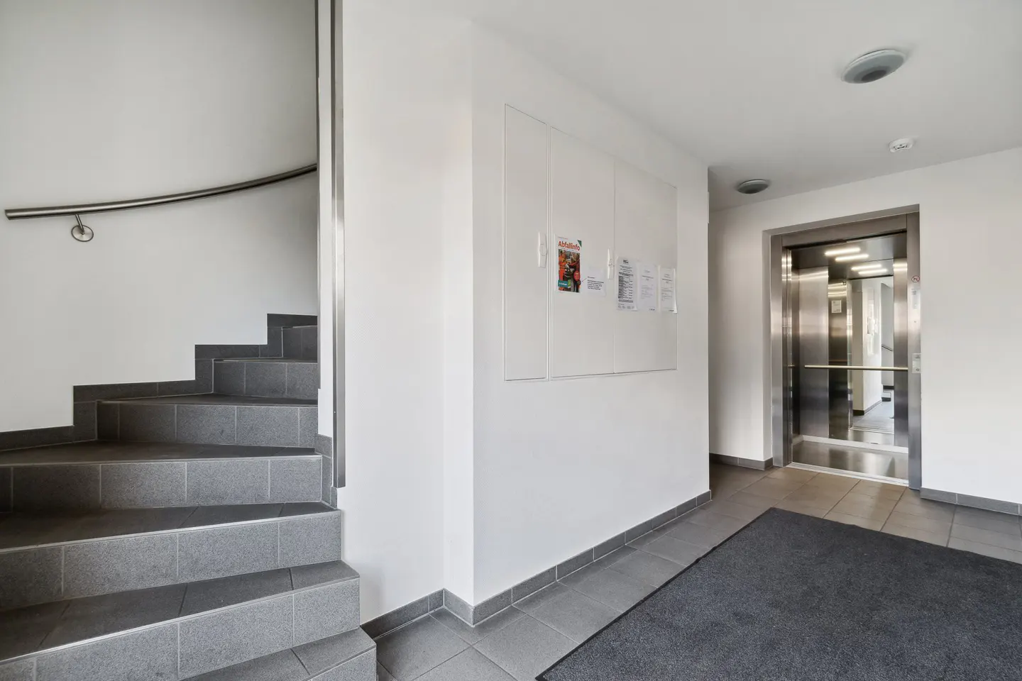Interior view of a building lobby with gray stairs, a stainless steel elevator, and a bulletin board on a white wall.