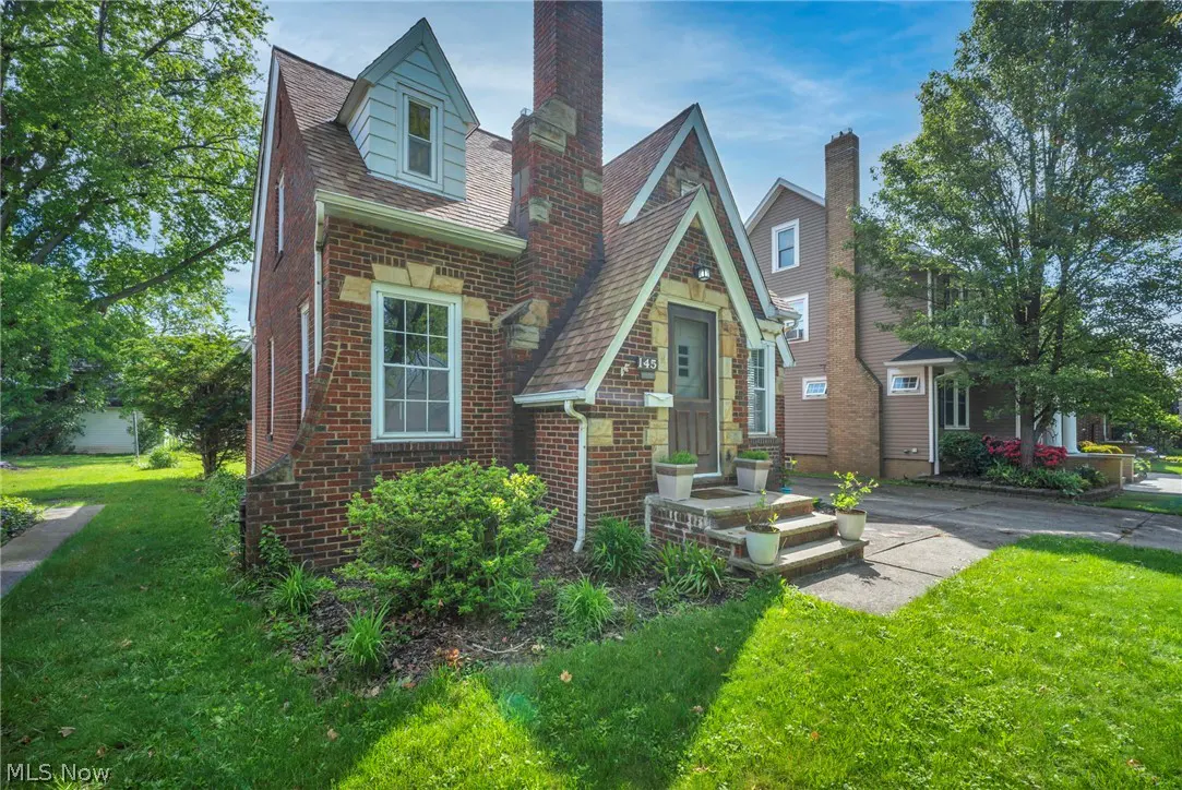 A two-story brick house with a chimney and a peaked roof, surrounded by green grass and trees.