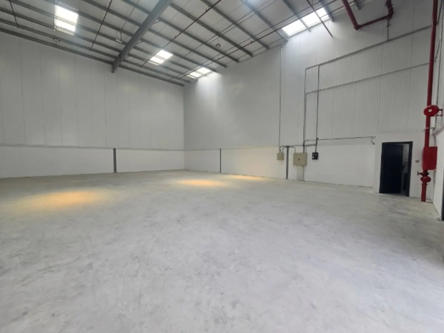 Empty warehouse interior with white walls, concrete floor, and skylights. Red fire suppression system visible.