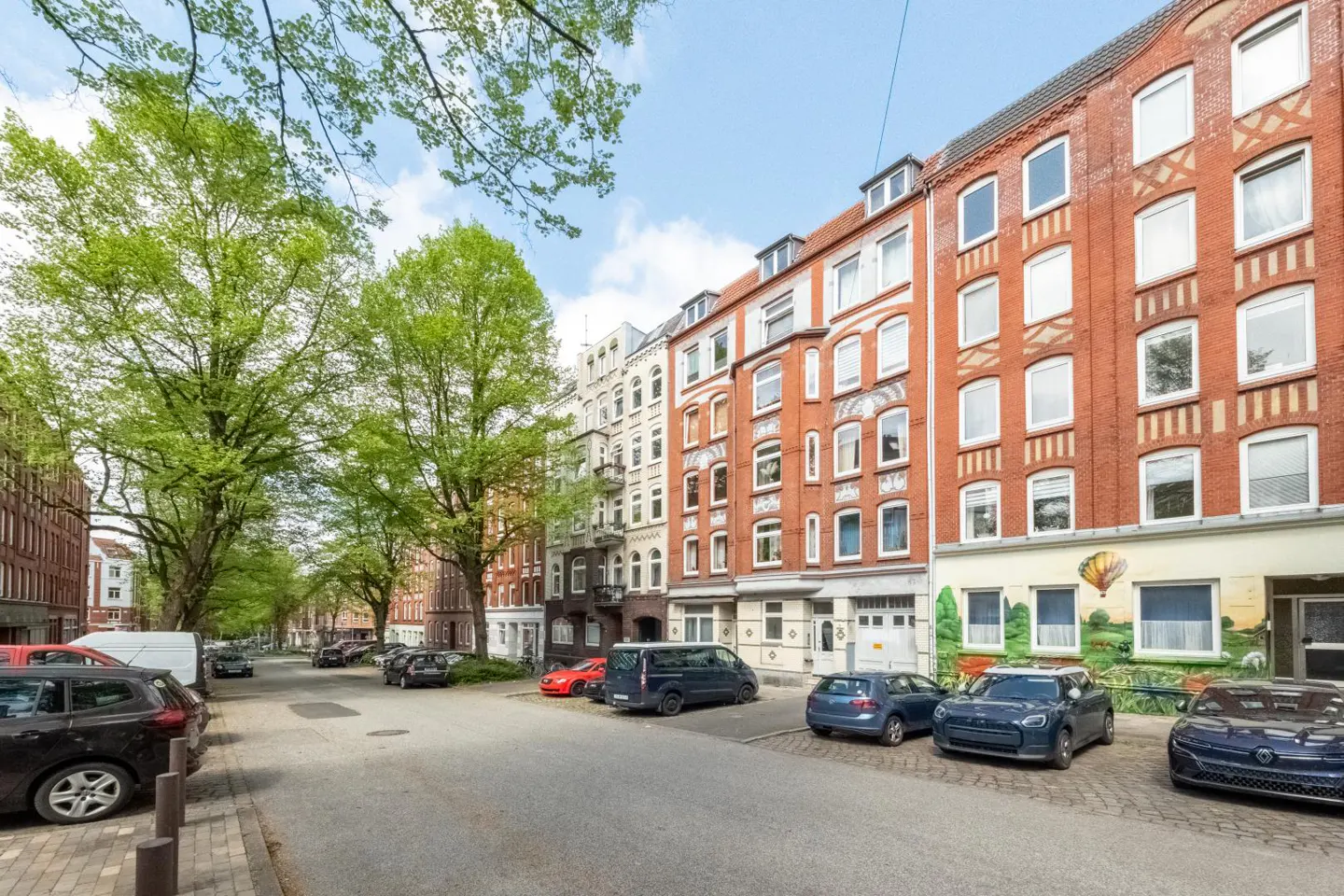 Street view of a row of historic red brick apartment buildings with parked cars and lush green trees on a sunny day.