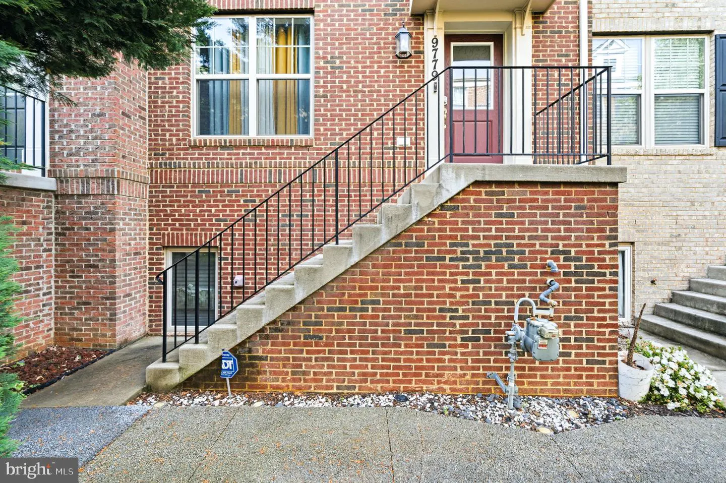 Exterior view of a brick townhouse with concrete stairs leading to a red front door with black railings.