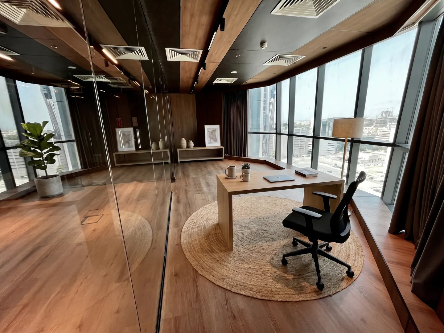 A modern office space with a desk, chair, and large windows offering a city view. The room features wood accents and a neutral color palette.