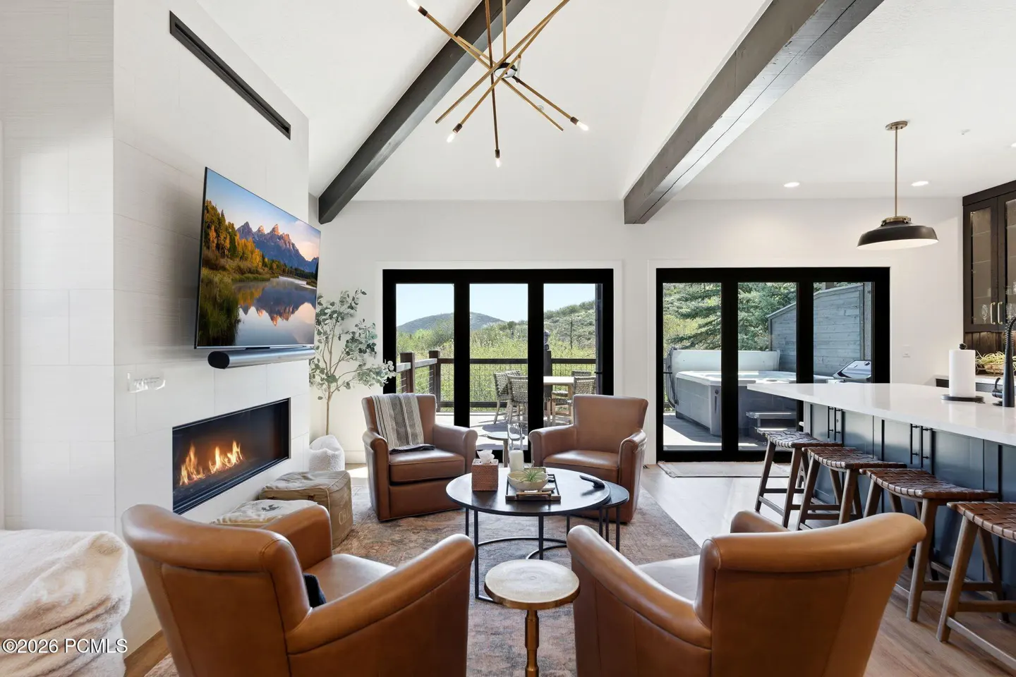 Bright living room with vaulted ceiling, beams, and leather chairs around a coffee table. Fireplace, TV, and mountain view through black framed doors.
