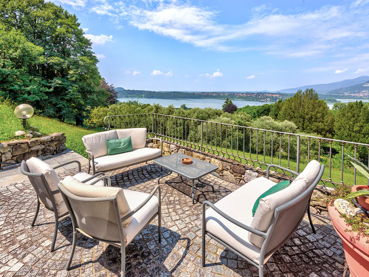 Outdoor patio with white cushioned seating, a metal table, and a stone floor overlooking a lake and green landscape under a blue sky.