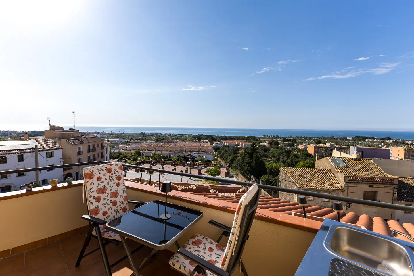 Balcony view with table, chairs, and sink overlooking a town and the sea under a clear blue sky.