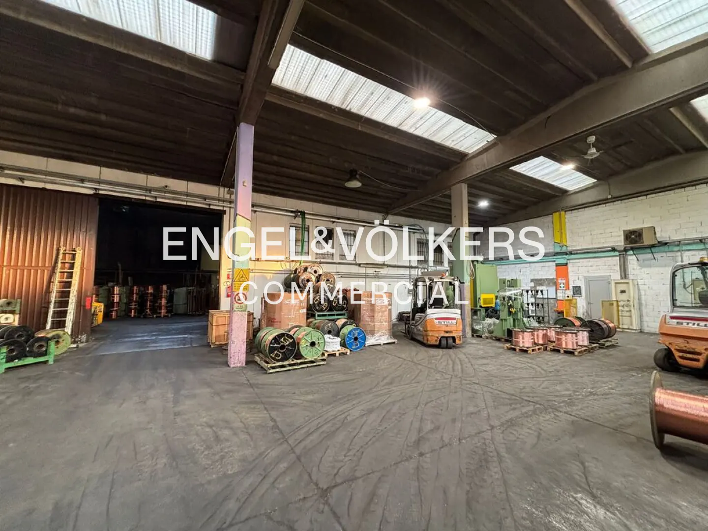 A wide shot of a warehouse with forklifts, machinery, and spools of wire. The Engel & Volkers logo is superimposed in the center.