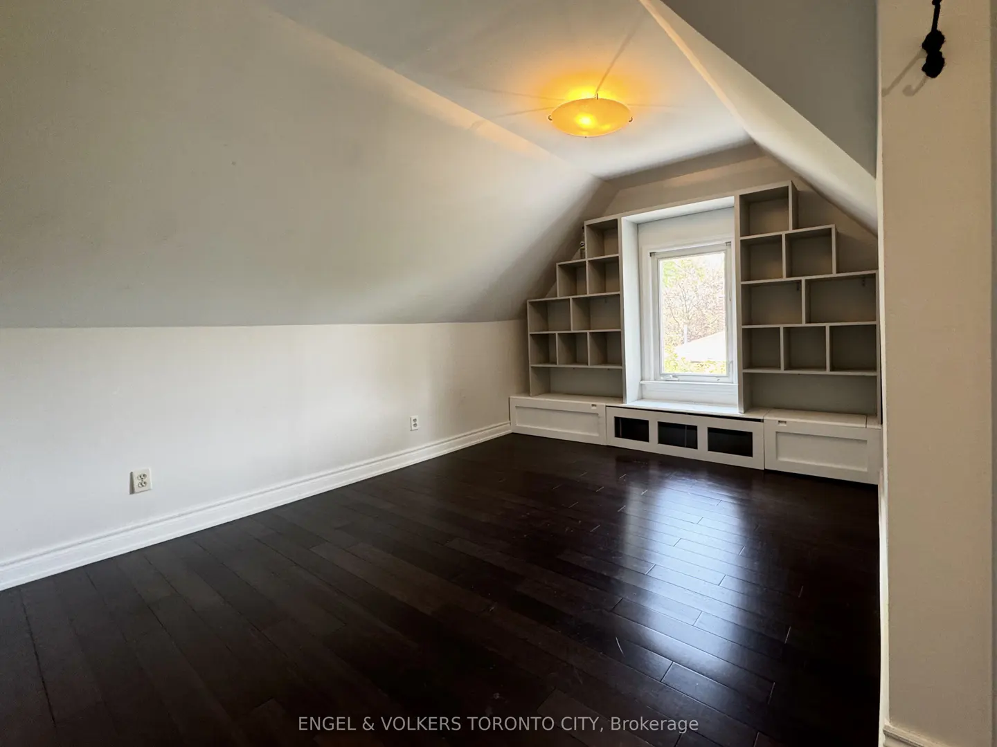 Attic room with dark wood floors, white walls, and built-in shelves around a window. A ceiling light fixture is visible.