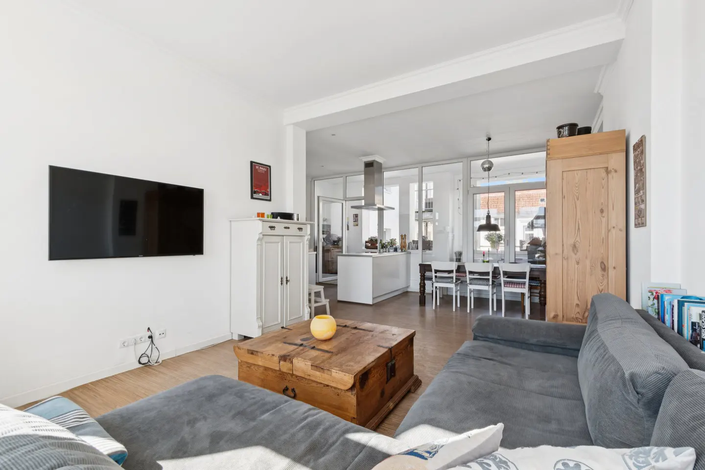 Living room with a grey sectional sofa, rustic wooden coffee table, wall-mounted TV, and an open-plan kitchen in the back.