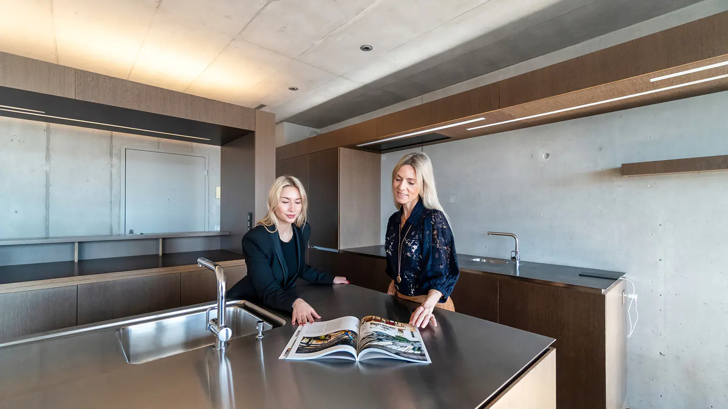 Two women looking at a magazine in a modern kitchen with stainless steel countertops and concrete walls.