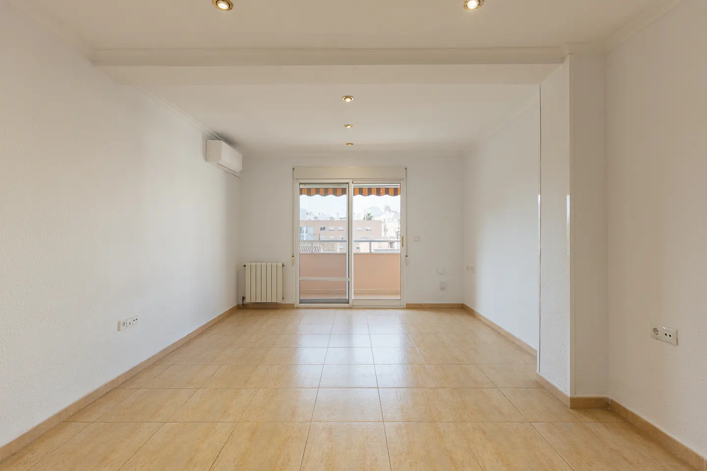 Bright, empty room with white walls, tan tile floor, and sliding glass doors to a balcony with a city view.