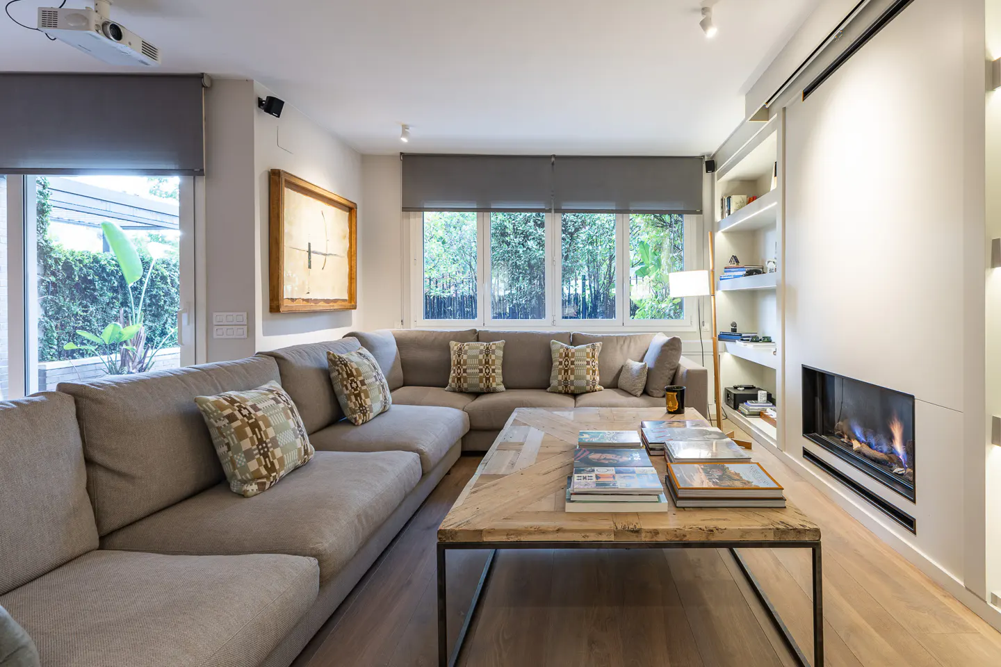 Bright living room with a large gray sectional sofa, a rustic wood coffee table, and a modern fireplace. Large windows offer a view of lush greenery.