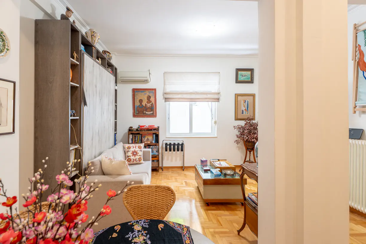 A bright living room with a sofa, bookshelf, coffee table, and parquet floor. Red flowers are in the foreground.
