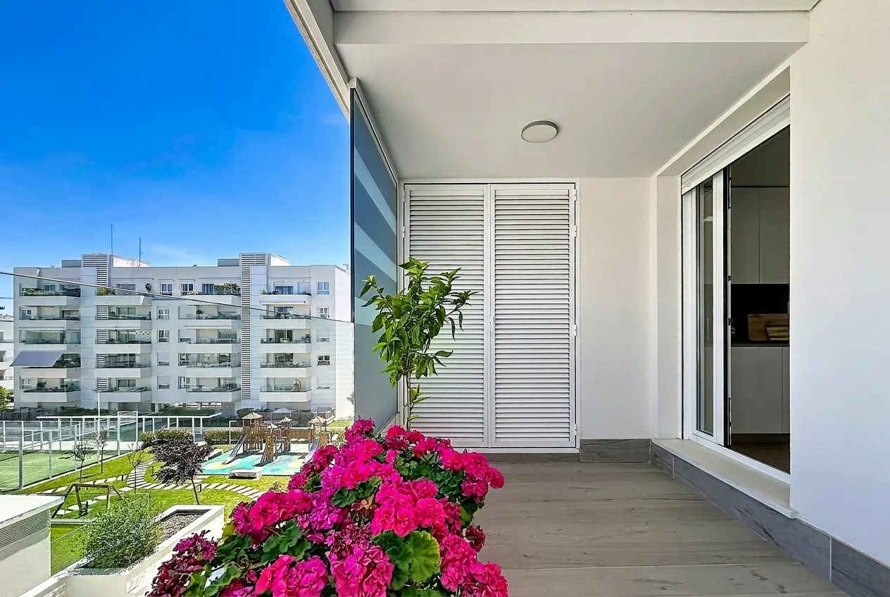 Balcony view with pink geraniums, a small tree, and a playground below. White buildings are visible in the background under a blue sky.