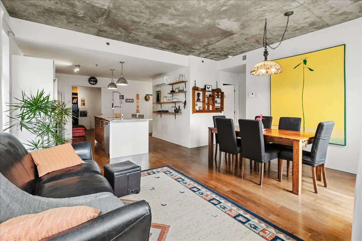 Open-concept living space with a black leather sofa, wood floors, and a dining table with black chairs. A yellow painting hangs on the wall.