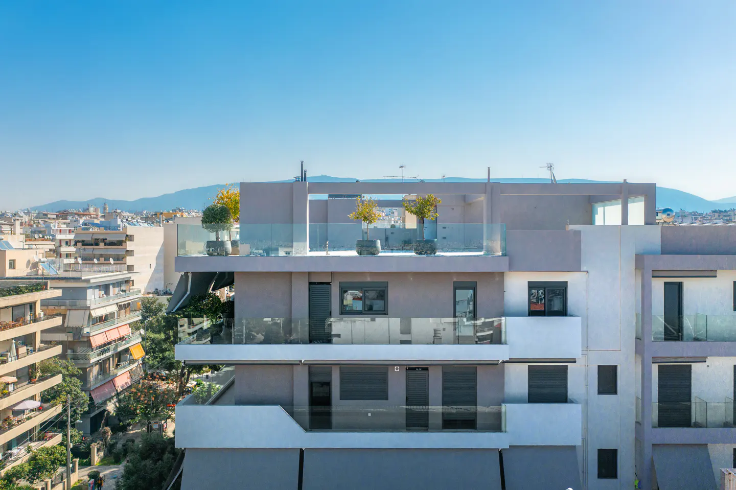 Modern apartment building with a rooftop terrace, glass railings, and potted trees against a clear blue sky.