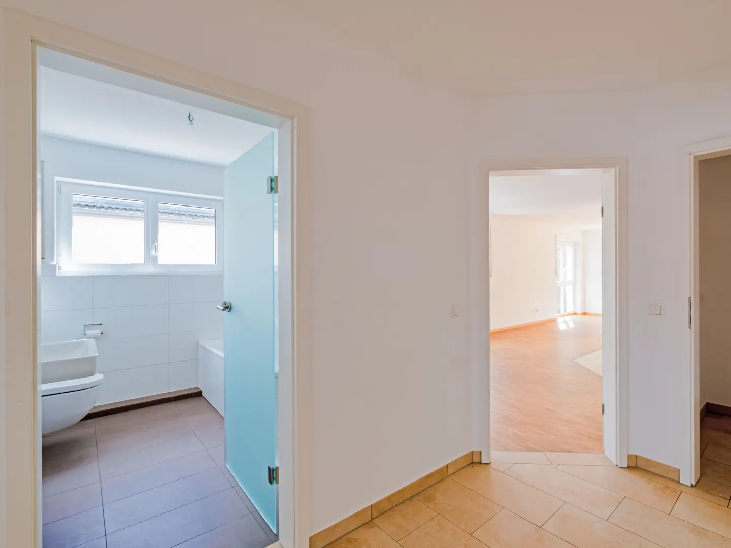 Hallway with tile floor and white walls. Open doors lead to a bathroom and a living room with hardwood floors.