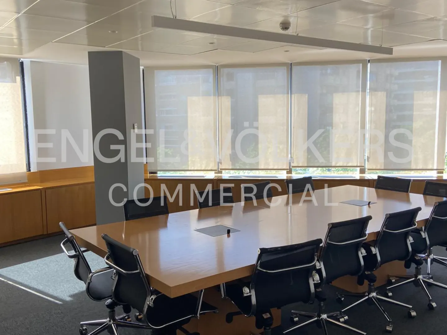 Bright office space with a large wooden conference table surrounded by black chairs, under a long light fixture.