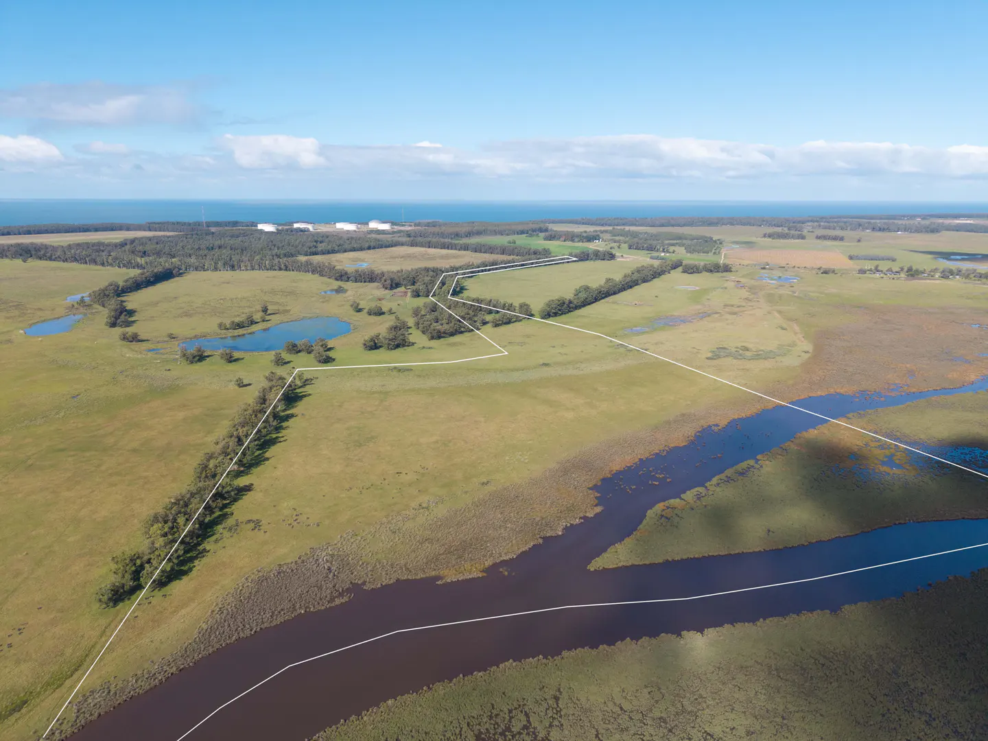 Aerial view of a large, green, undeveloped property with a river and ponds, outlined in white. Ocean and sky in the background.