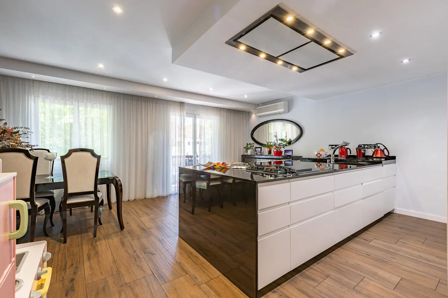 Bright, modern kitchen with a black island, white cabinets, and wood floors. A dining table sits near sheer curtains.