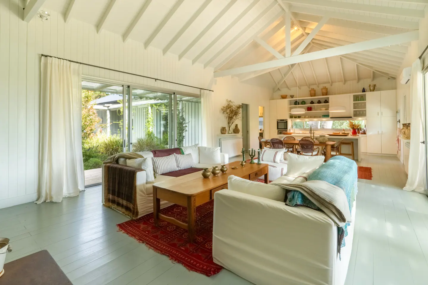 Bright, open-plan living room with white sofas, wooden table, and red rug. Kitchen and garden view in the background.