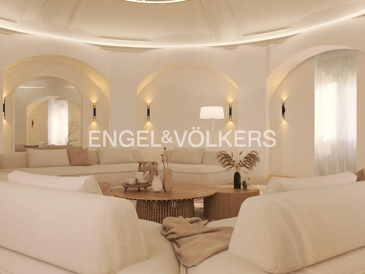 Bright living room with white sofas, arched doorways, and recessed lighting. A round wooden coffee table sits in the center. Engel & Völkers logo visible.