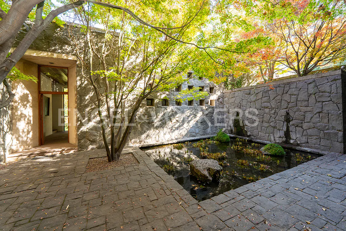 Stone courtyard with a pond, trees, and a building with an open doorway. The pond has rocks and moss. Trees have green and red leaves.