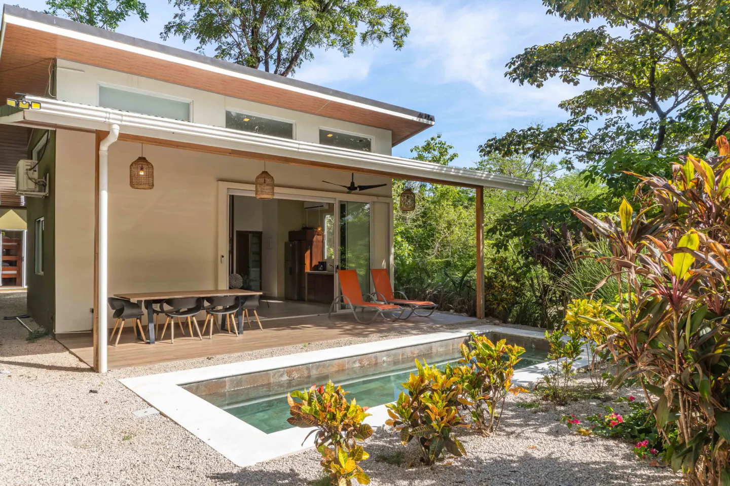 Exterior view of a modern home with a small pool, patio with table and chairs, and orange lounge chairs. Lush greenery surrounds the property.