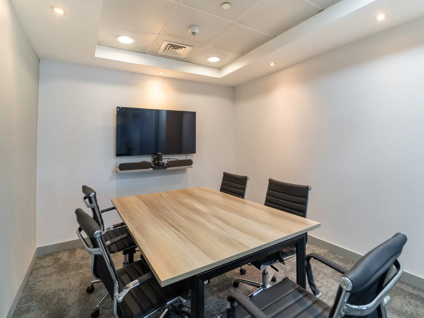A small conference room with a wood table, black chairs, and a wall-mounted TV. The walls are white, and the floor is carpeted.