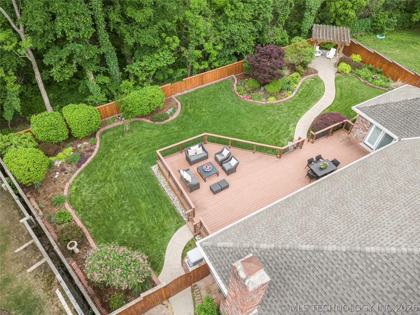 Aerial view of a home's backyard with a deck, patio furniture, green lawn, garden, and wooden fence.