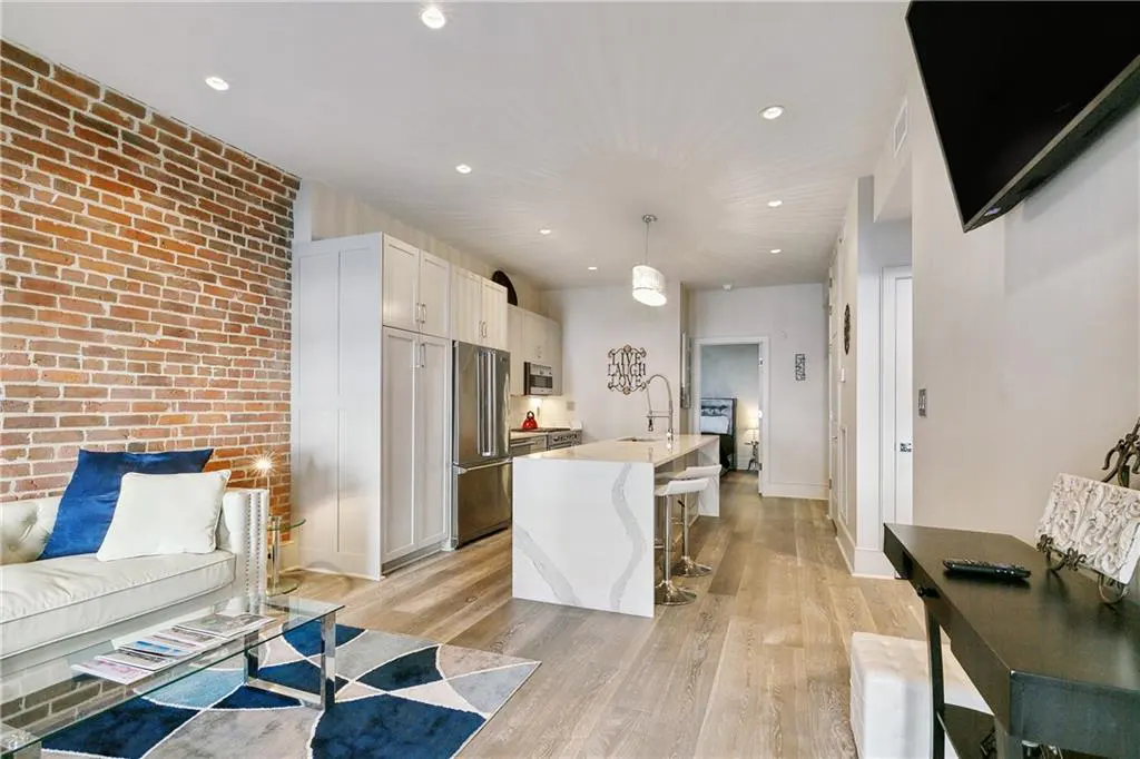 Open-concept apartment with brick wall, white cabinets, and island with bar stools. Blue and gray rug on wood floor.