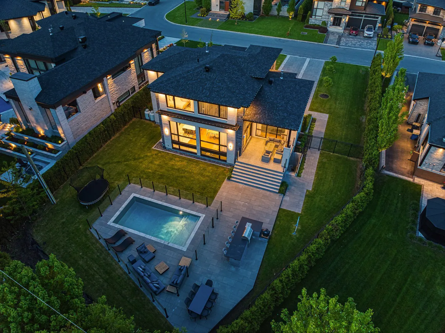 Aerial view of a modern home with a pool, patio furniture, and a dark roof. The house has large windows and is surrounded by green grass and trees.