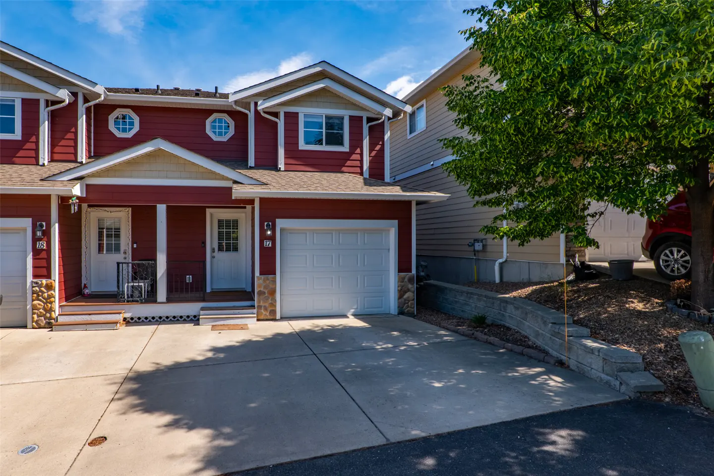 Exterior view of a red townhouse with a white garage door, concrete driveway, and a large tree on a sunny day.