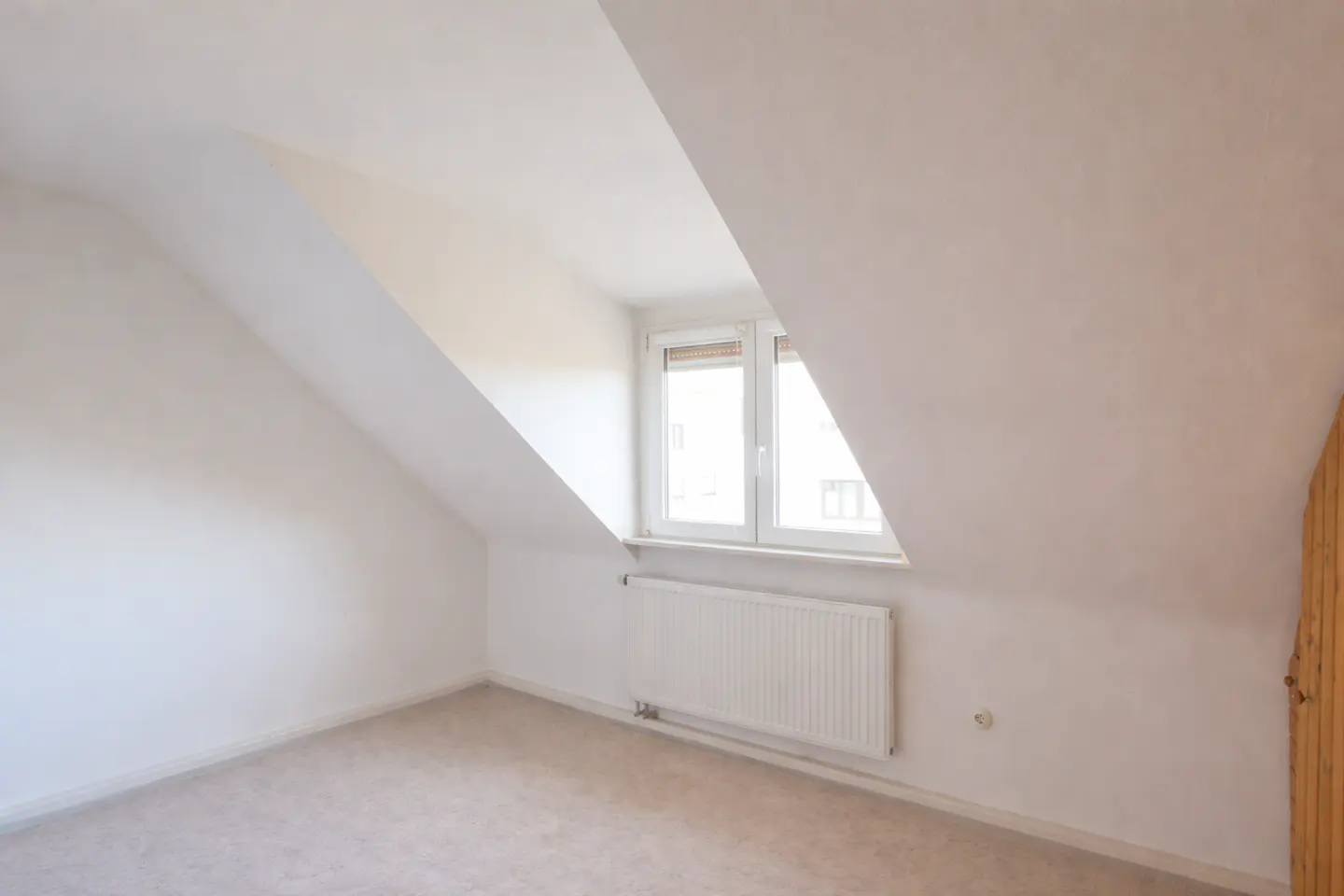 Attic room with white walls, beige carpet, and a window. A white radiator is under the window. A wooden door is on the right.