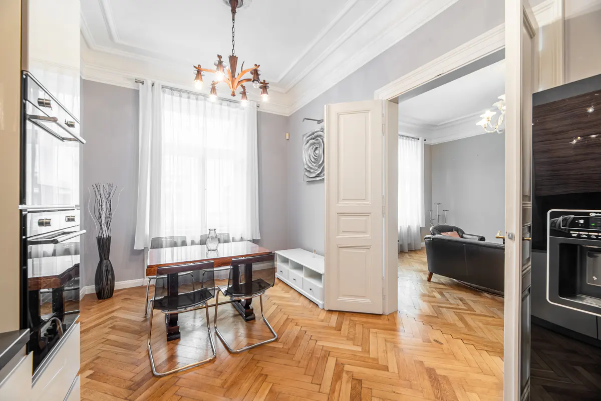 A dining room with wood floors, a table, chairs, and a chandelier. A doorway leads to a living room with a sofa.