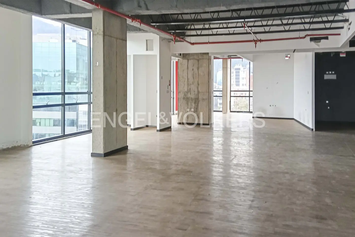 Empty office space with concrete pillars, large windows, and a polished concrete floor. Red pipes run along the ceiling. City view visible through the windows.