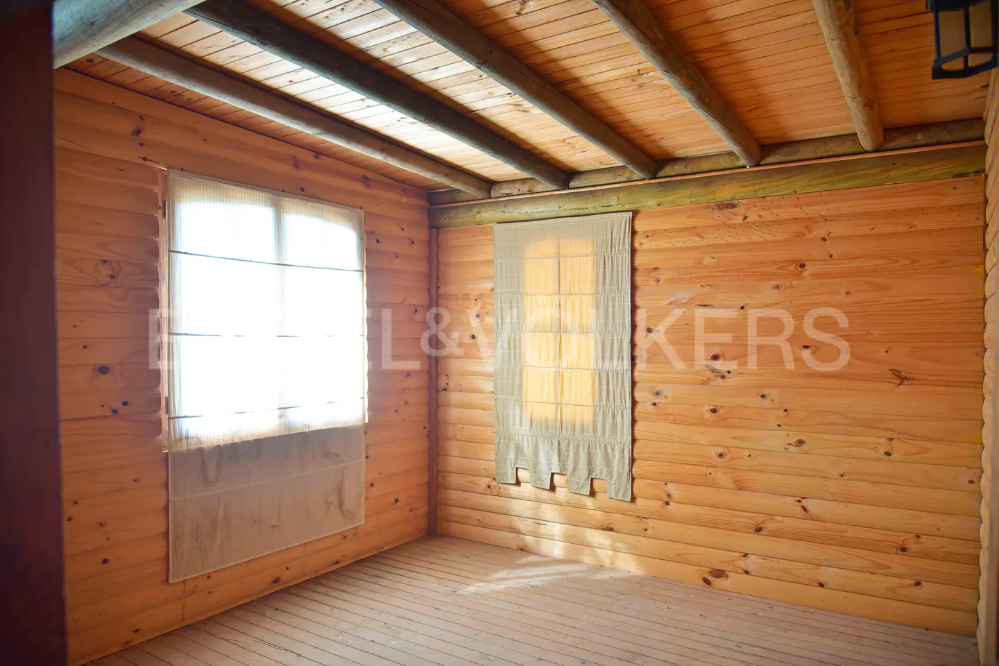 Empty room with wood walls, ceiling, and floor. Two windows with beige curtains.