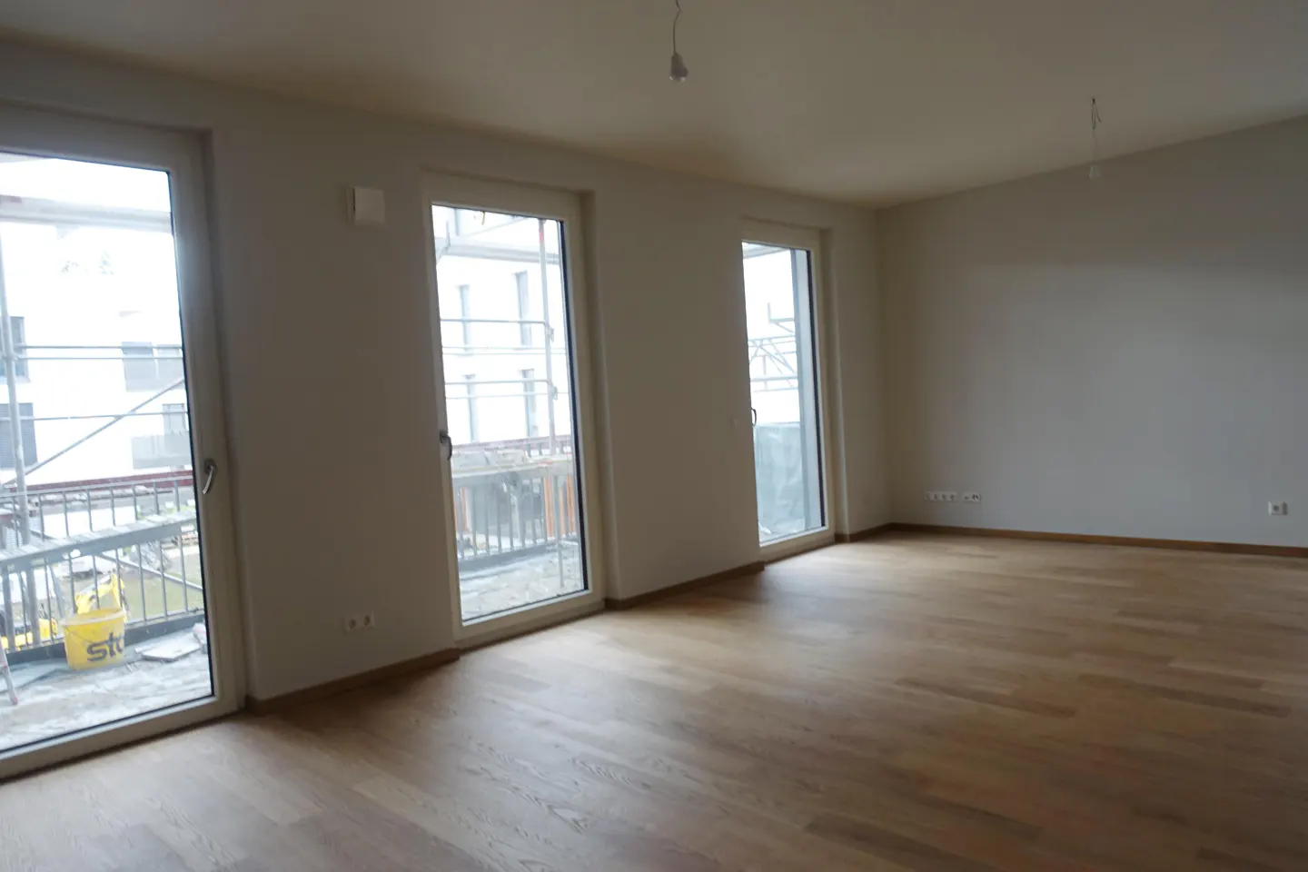 Unfurnished room with light gray walls and wood floors. Three white-framed glass doors lead to balconies.