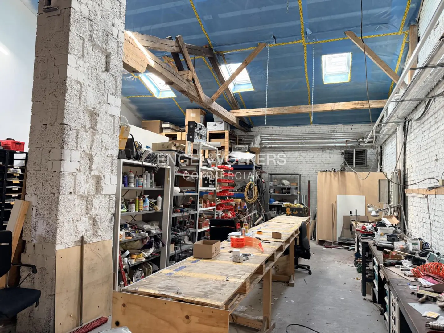 Interior of a commercial space with exposed brick, wooden beams, and skylights. Shelves and workbenches are filled with tools and supplies.