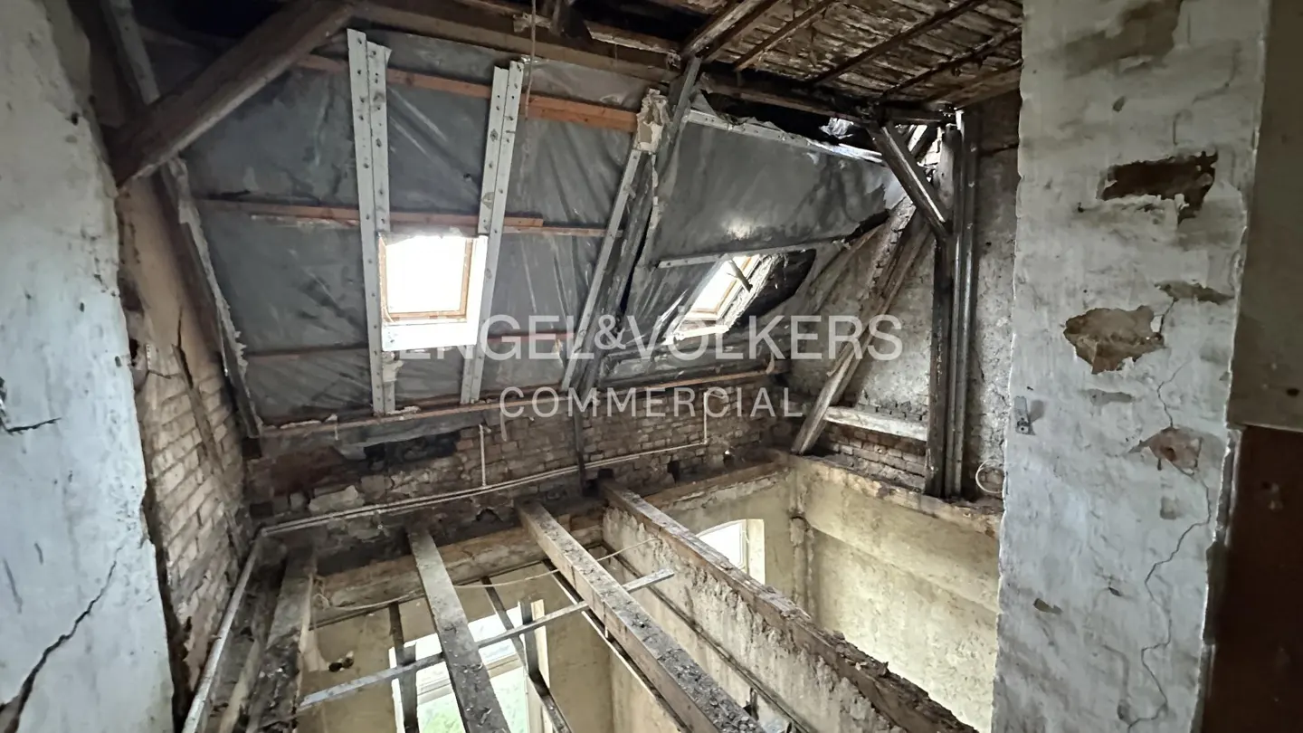 Interior view of a dilapidated attic with exposed beams, brickwork, and skylights. Walls are cracked and peeling, showing signs of disrepair.
