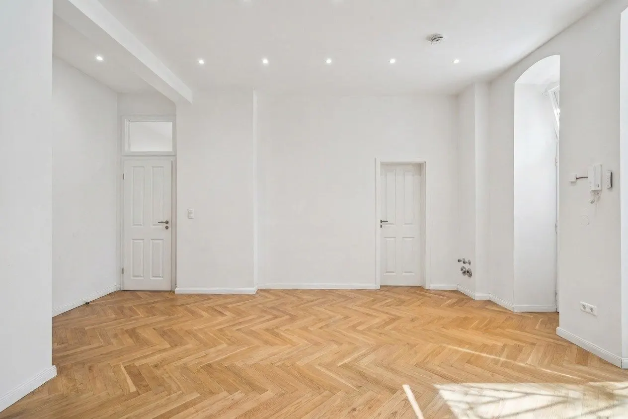 Bright, empty room with white walls, herringbone wood floor, and two white doors. Sunlight shines in from a window on the right.