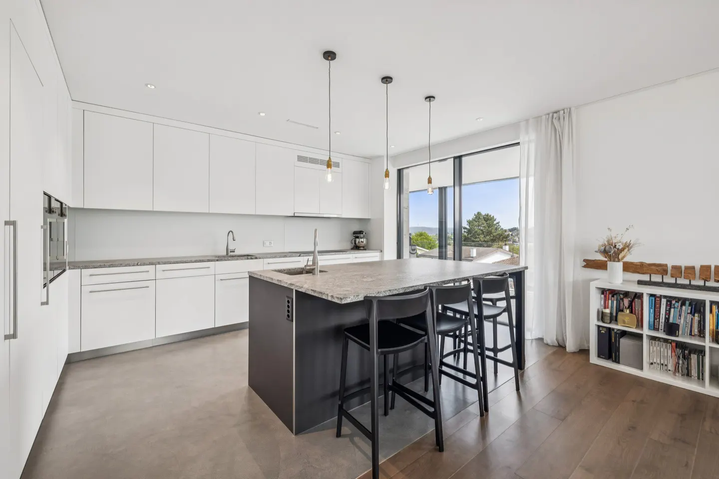Bright kitchen with white cabinets, a dark island with a granite countertop, and black stools. A large window offers a view of the outdoors.
