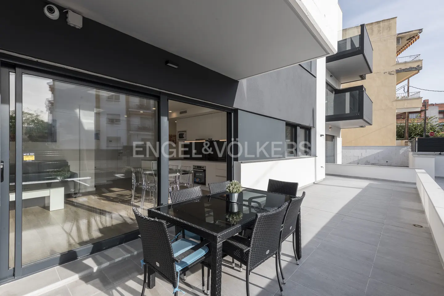 Outdoor patio with black table and chairs. Sliding glass doors lead to a modern kitchen. Engel & Volkers logo on the glass.