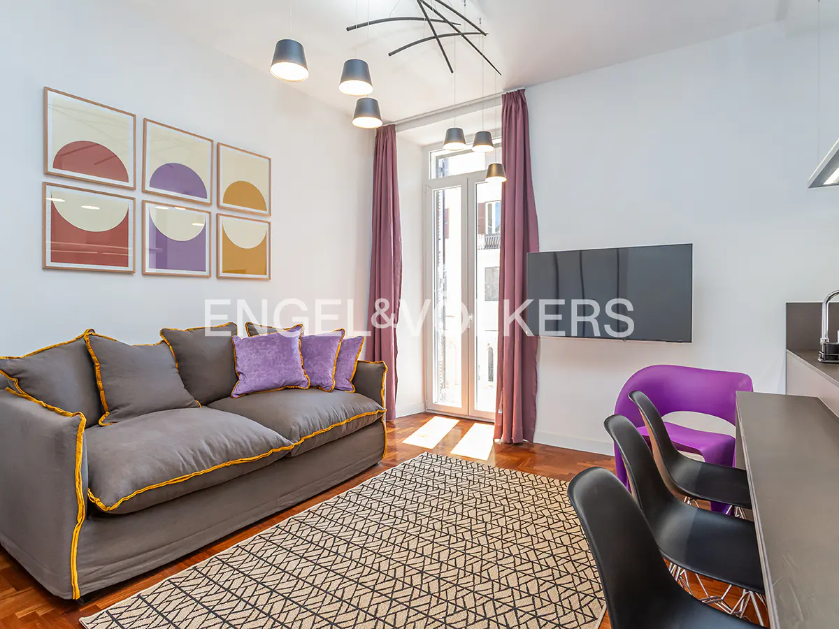 Living room with gray sofa, purple pillows, geometric rug, and modern art. Natural light streams through the balcony door.