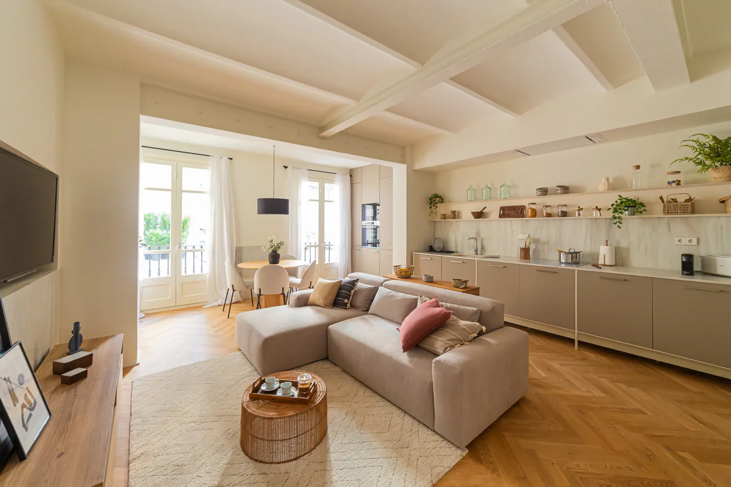 Bright, open-concept apartment with a beige sectional sofa, round wicker table, and a modern kitchen with light gray cabinets. French doors lead to a balcony.