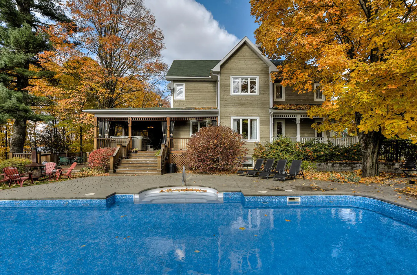 A two-story house with a green roof and siding, a blue pool, and fall foliage.