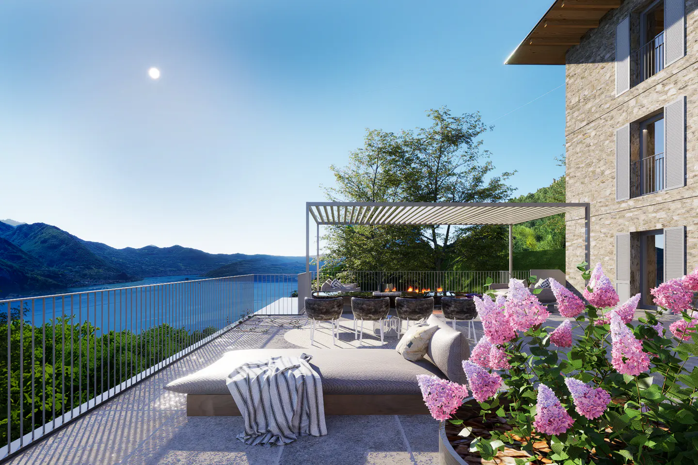 Outdoor patio with lake view. Lounge seating, dining table under pergola, and pink flowers. Stone building on right.