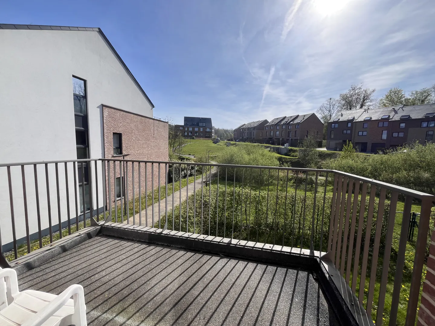 Balcony view with metal railing overlooking green space and houses under a blue sky.