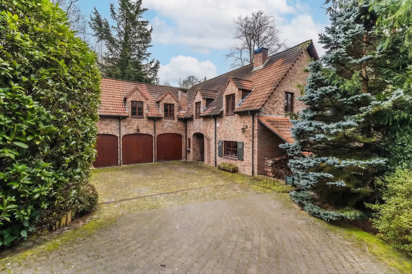 Exterior view of a brick house with a red tile roof, three-car garage, and a paved driveway surrounded by green trees and bushes.