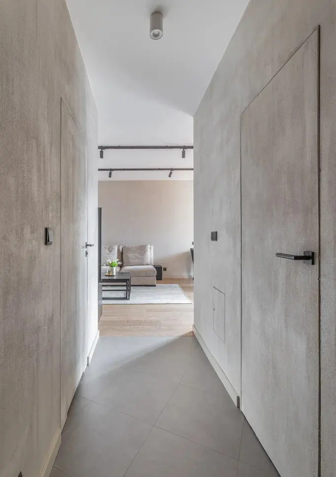Hallway view with gray walls, doors, and floor tiles. A living room with a beige sofa and black coffee table is visible in the background.